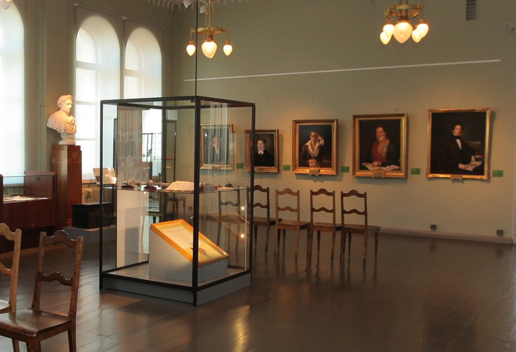 A colour photo from a museum exhibition, with a display case in the middle. At the bottom is a framed document placed on a slanted platform. In the background are portraits and wooden chairs. On the left, by some arched windows, is a marble bust on a stand.