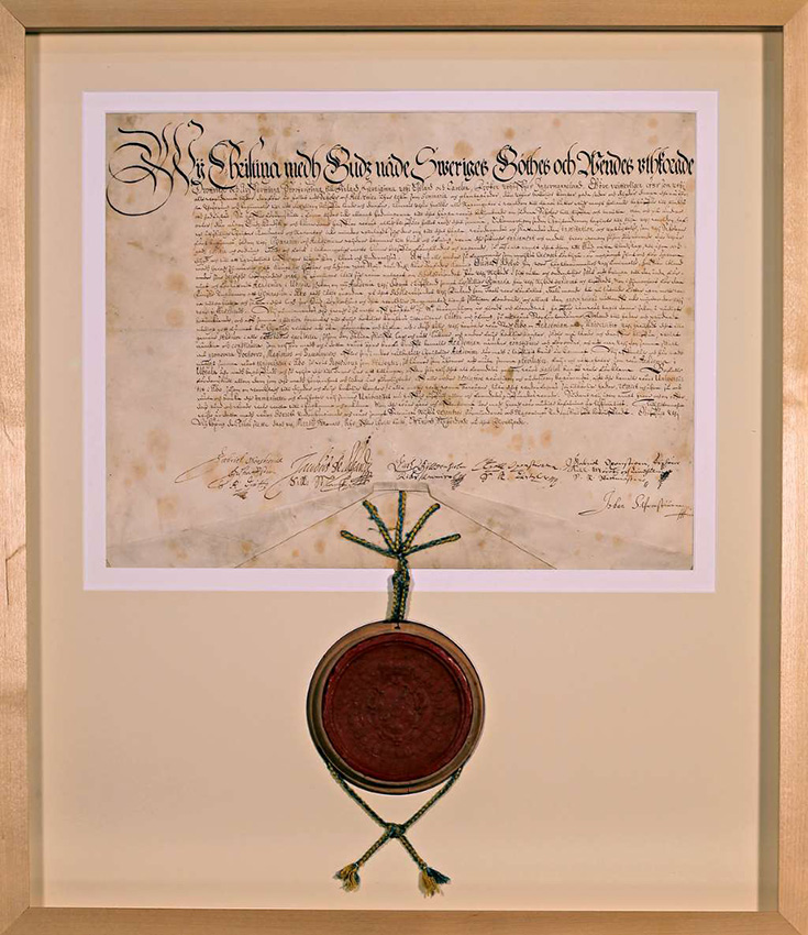 A framed letter written in elaborate handwriting, with a round red wax seal at the bottom. The letter is surrounded by light-coloured matting and a pale, narrow and smooth wooden frame.