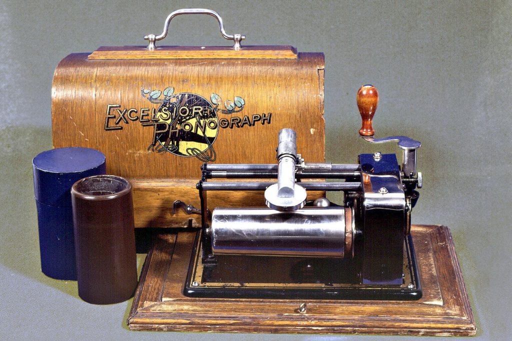 : A metal device with a cylinder and a crank, on a wooden base. Next to the device is a brown wax cylinder and, in the background, the wooden lid of the device, with a metal handle.