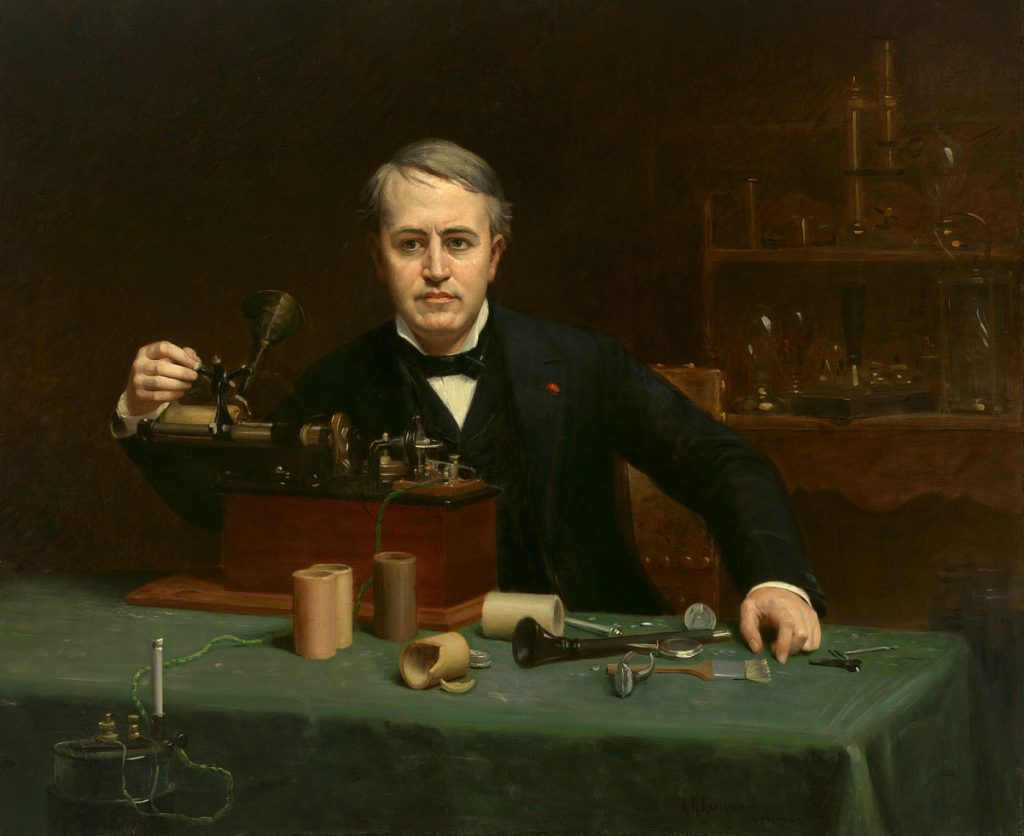 A brown-haired man in a black jacket and a white shirt sits at a table, using or building a device with metal parts on a wooden base. Cylinders and other components of the device are also lying on the table, covered by a dark green tablecloth.