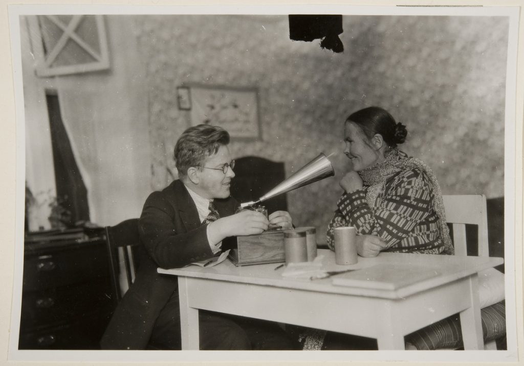 A man with glasses and a woman with her hair tied back are sitting at a white table. The latter is talking into the horn of a device on the table. Also on the table are cylinders belonging to the device.