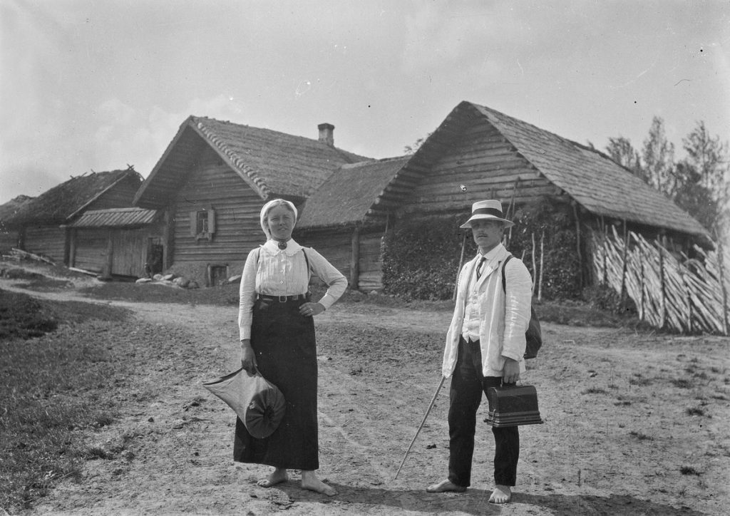 A man and a woman standing in front of log buildings. The man is holding a phonograph in its case, and the woman has a phonograph horn in a cloth bag.