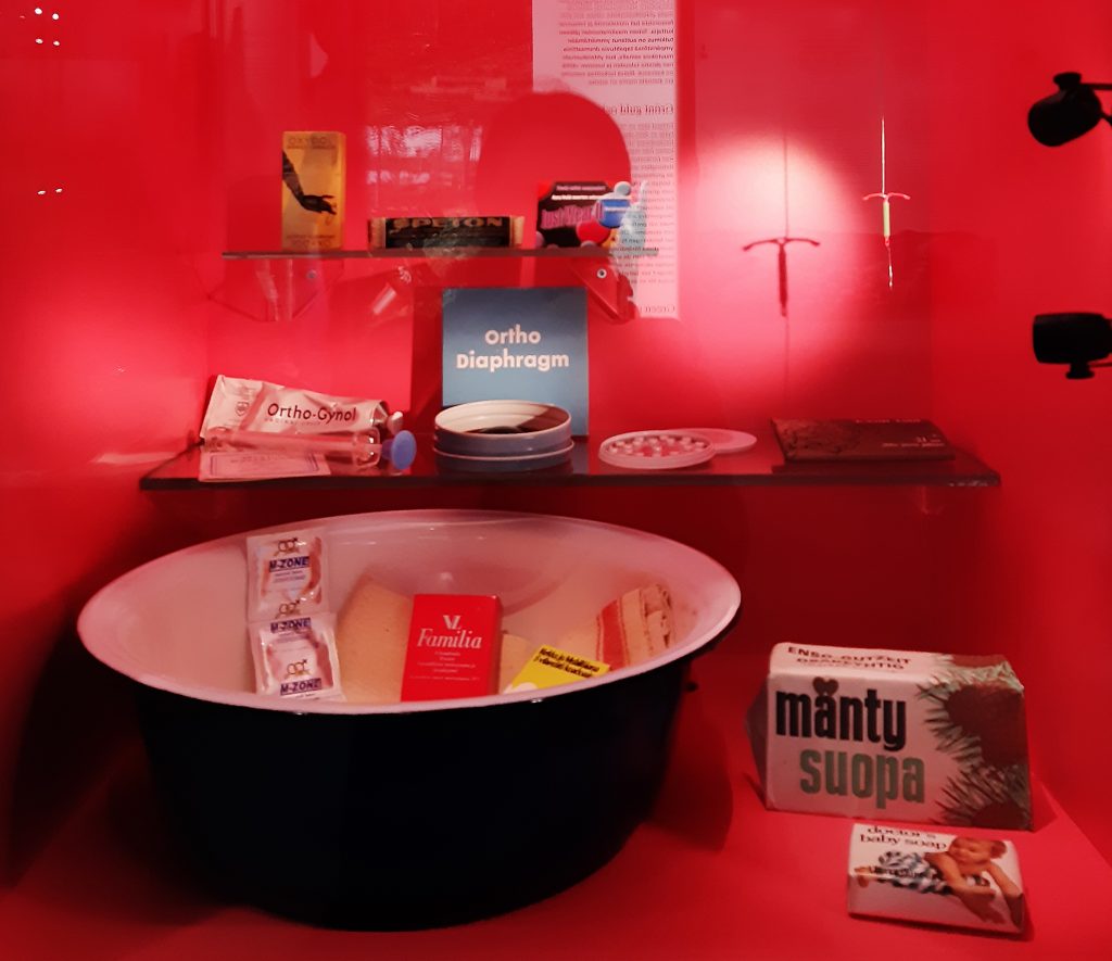 The photo shows a small square display case, with a raspberry-red interior and a glass front, that contains various contraceptives and maternity package objects, placed on three different levels.