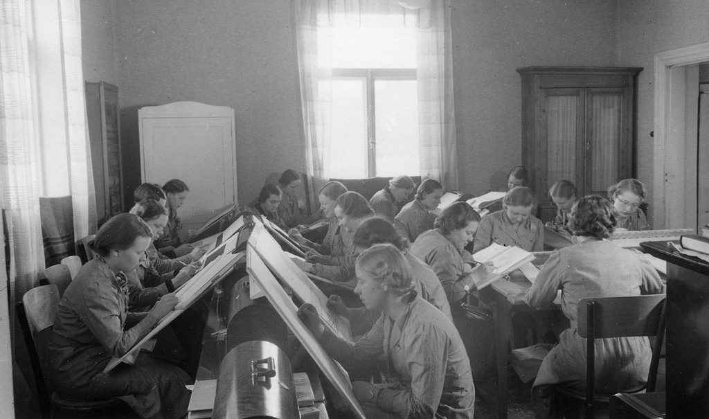 A large group of women students in a smallish classroom drawing or writing on large sheets of paper or into workbooks placed on inclined side tables. The students are wearing uniform outfits. There are two windows letting in light and two cabinets in the room along the back wall.
