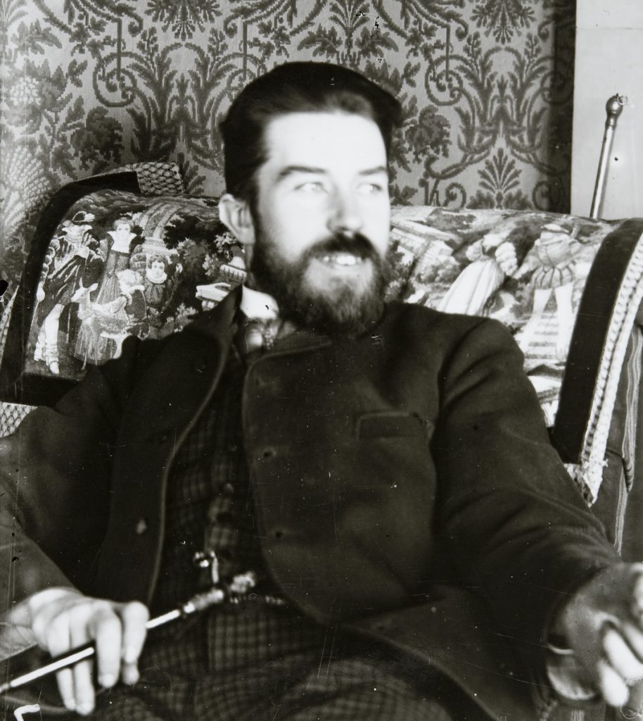 A black-and-white photograph of a bearded man in an armchair. In the background is a wallpaper of floral design.