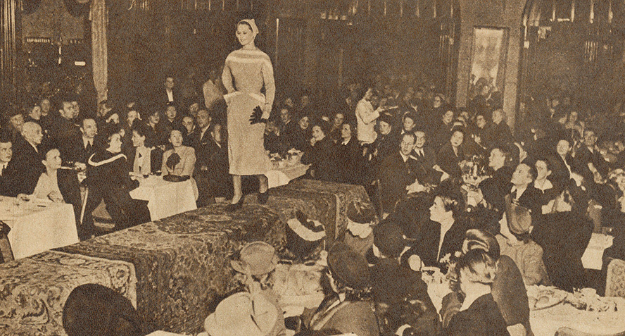 Mustavalkoinen kuva muotinäytöksestä, istuva yleisö, jonka keskellä catwolilla kävelevä malli.