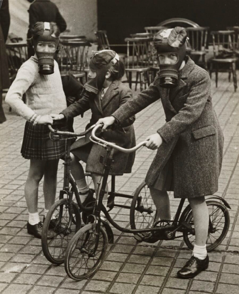 Kolme pientä tyttöä kadulla kaasunaamarit päässään. Kahdella tytöistä on polkupyörä. Taustalla näkyy runsaasti puutuoleja. Kuva on mustavalkoinen.
