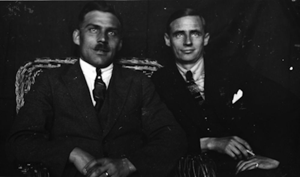Mustavalkoinen puolivartalovalokuva kahdesta tummaan pukuun ja solmioon pukeutuneesta miehestä, jotka istuvat rottinkisohvalla ja hymyilevät vienosti. Toisella on sytyttämätön tupakka sormien välissä.