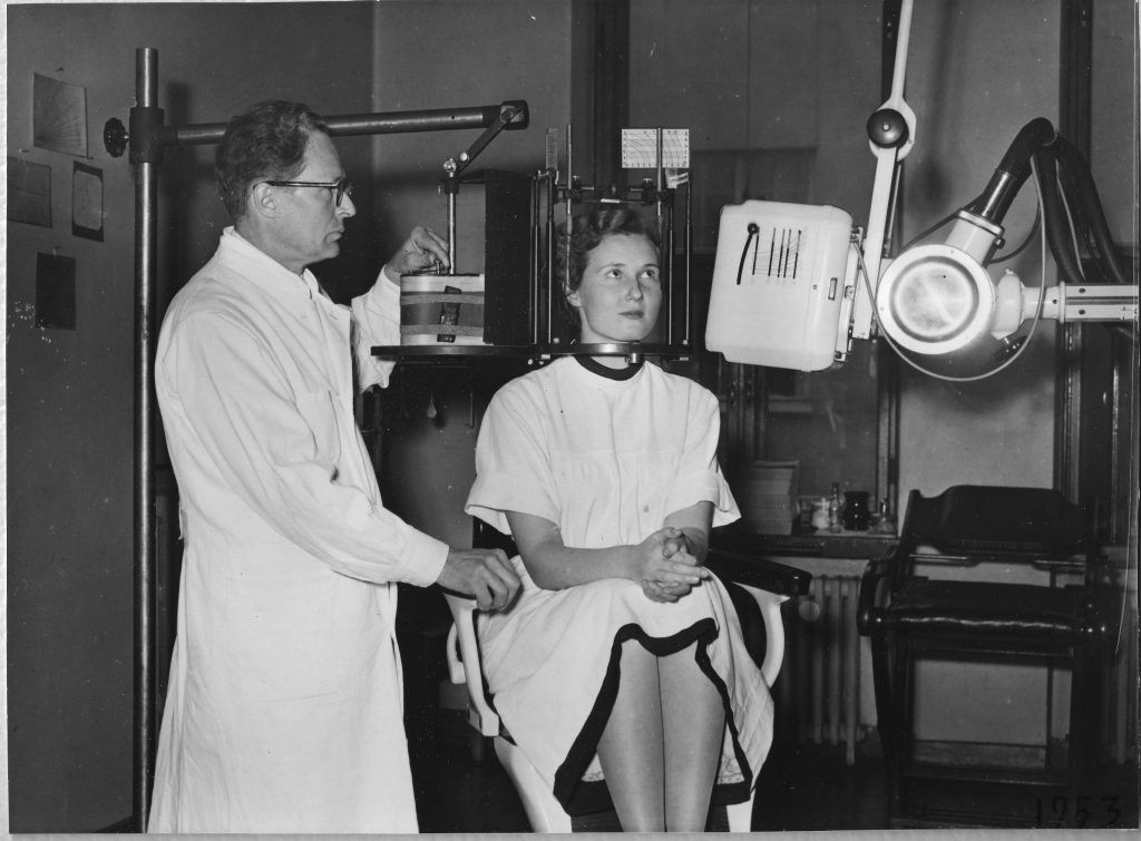 Tuolilla istuva nainen, jonka pään ympärillä on metallinen häkkyrä ja oikealla puolella röntgenkamera ja vasemmalla valkotakkinen mies, joka säätää telineeseen asetettua kaarevaa röntgenfilmilevyä.