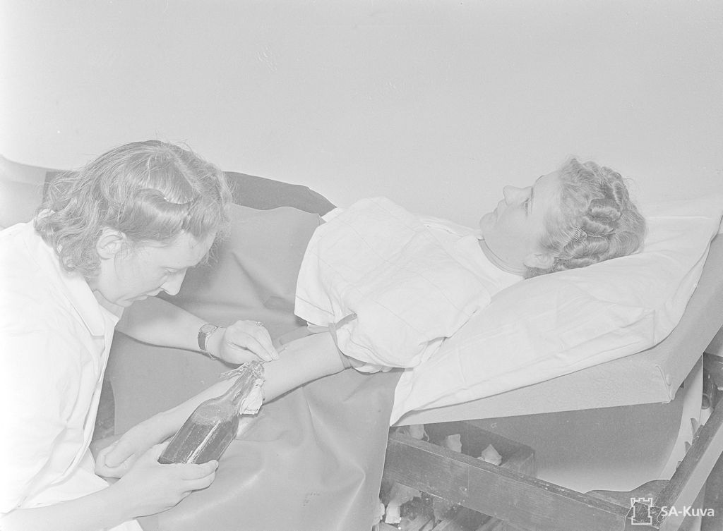 Nainen makaa tutkimuspöydällä. Hänen käsivarressaan on puristusside, ja toinen nainen yllään lääkärintakki valuttaa suuren neulan avulla verta lasipulloon. 