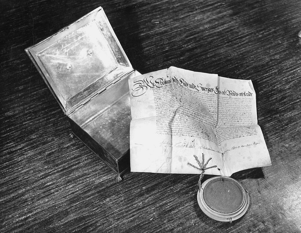 Mustavalkoinen kuva, jossa on avattu kiiltävä rasia ja sen oikean alakulman päälle ja pöydälle levitetty kirje taitoksineen ja pyöreä sinetti.