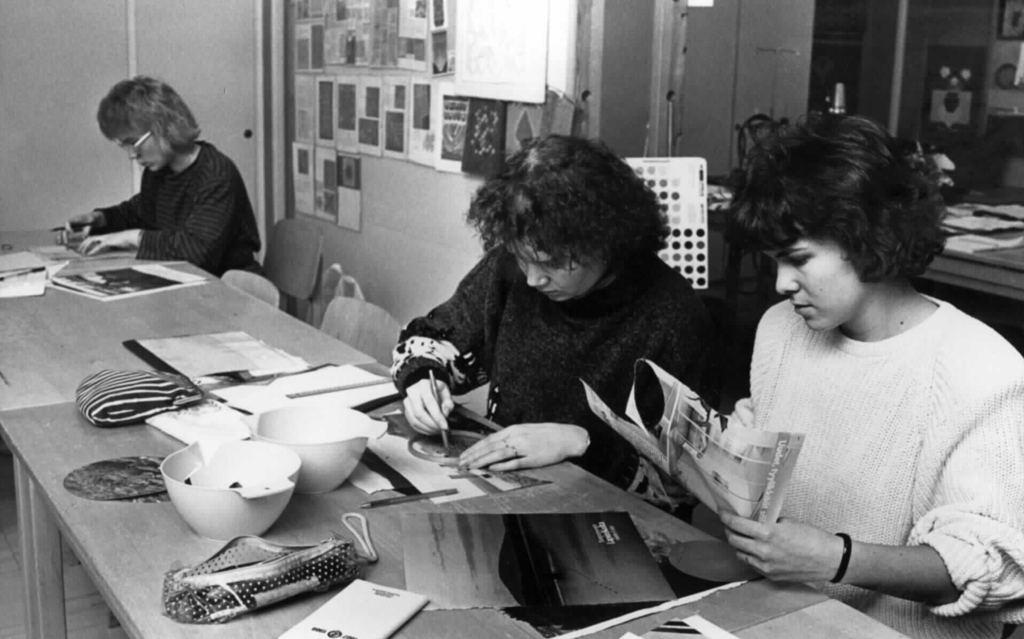 Kolme naispuolista opiskelijaa istuu pöydän ääressä ja tekee harjoitustehtävää, johon sisältyy lehtileikkeiden leikkausta.