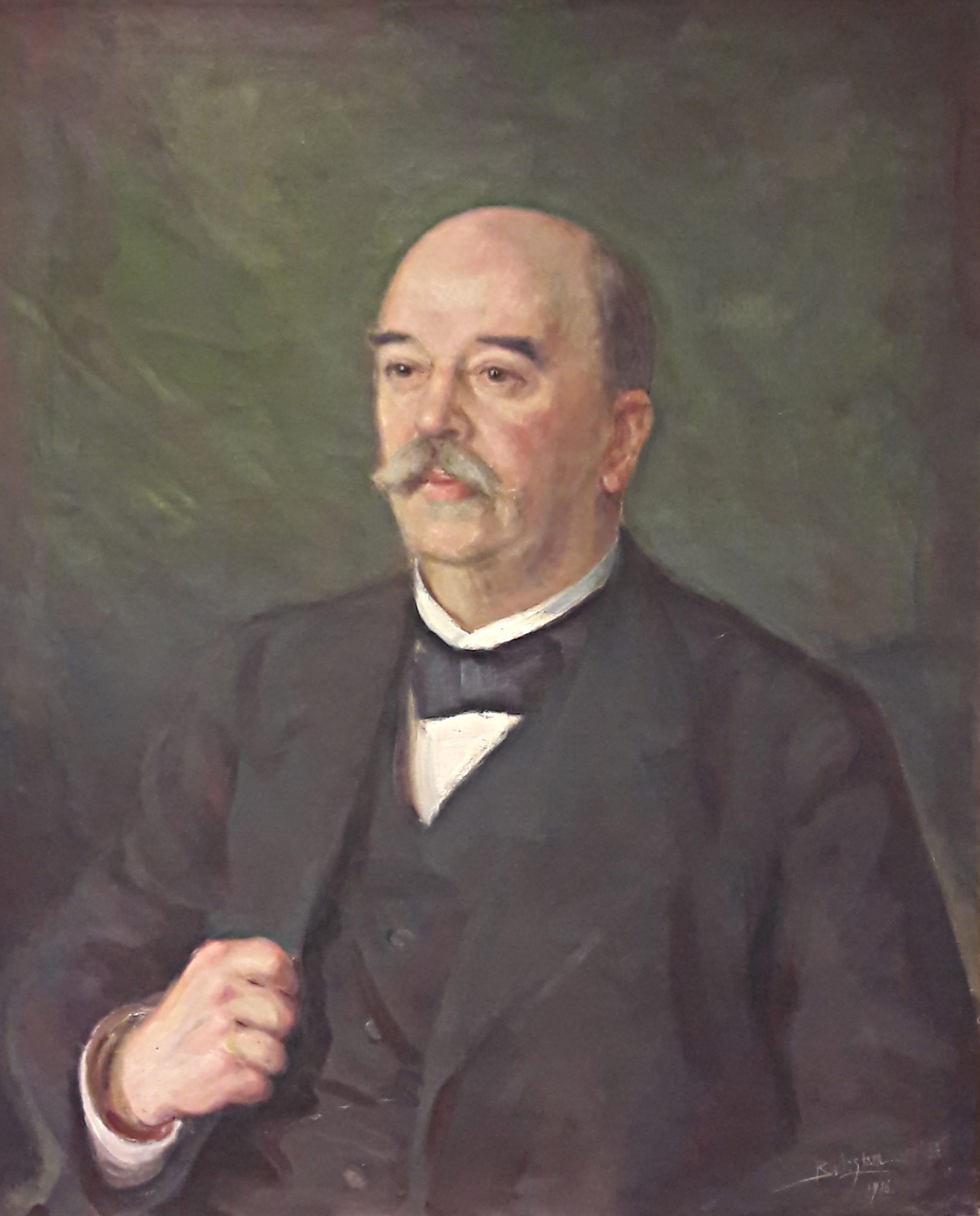 A half-body portrait of a man wearing a dark suit, white shirt and dark bow tie around his neck. He is holding the lapel of his suit jacket with one hand. He is partly bald and has a large moustache.