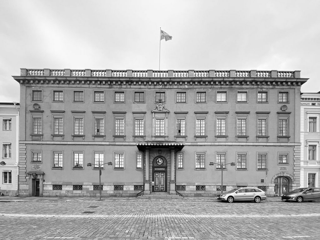 Mustavalkoisessa kuvassa näkyy kolmikerroksisen, matalan kivirakennuksen säännöllinen julkisivu ikkunoineen. Katolla liehuu Ruotsin lippu ja rakennuksen edustalle on pysäköity autoja.