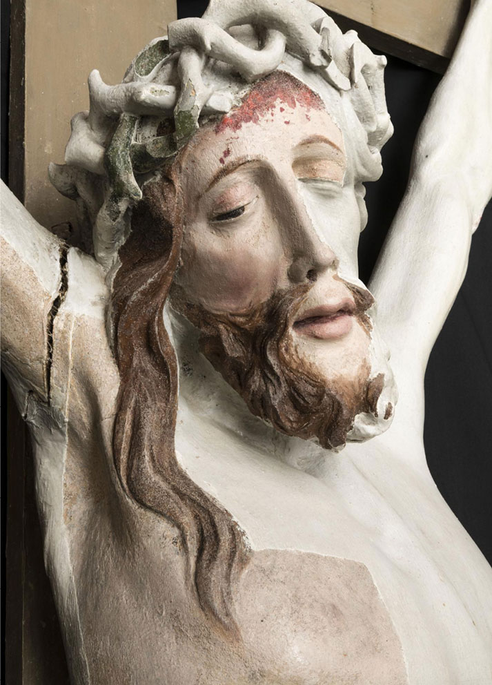 Lähikuva Kristus-veistoksen kasvoista ja rinnasta. Veistosta peittävä valkoinen maali on osittain poistettu niin, että kuvassa näkyy orjantappurakruunun vihreää väriä, verta otsalla, ruskea parta ja hiukset, sekä ihon väriä kasvoilla ja rinnalla.