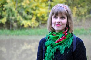 Photo of Emmi Kuittinen wearing a green scarf, smiling and looking into the camera, against a background of greenery