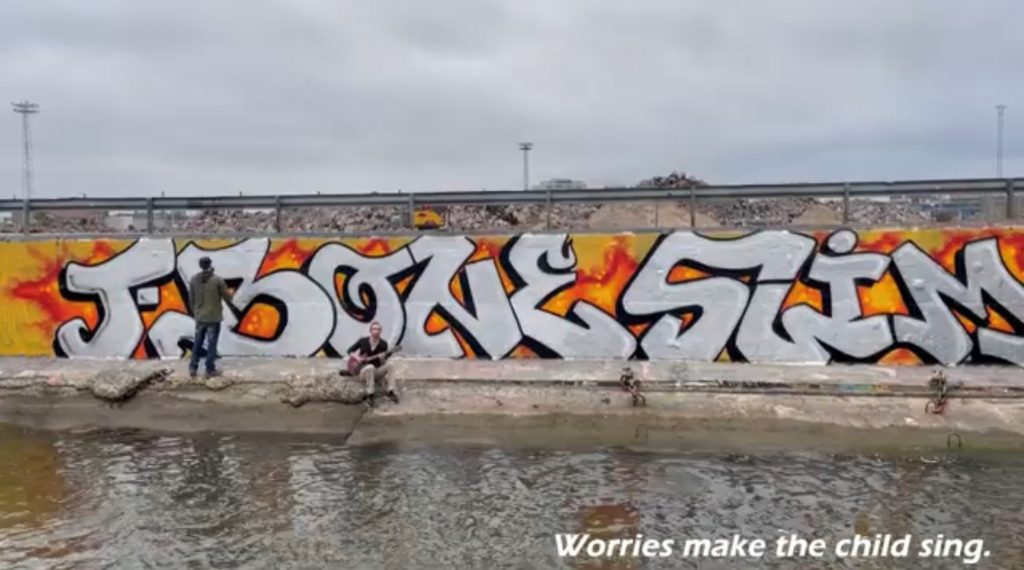 Graffiti “T-Bone Slim”. Two men: one standing, doing graffiti and other sitting and playing guitar. Water in front, road railing in the back. Text in the bottom: "Worries make the child sing."