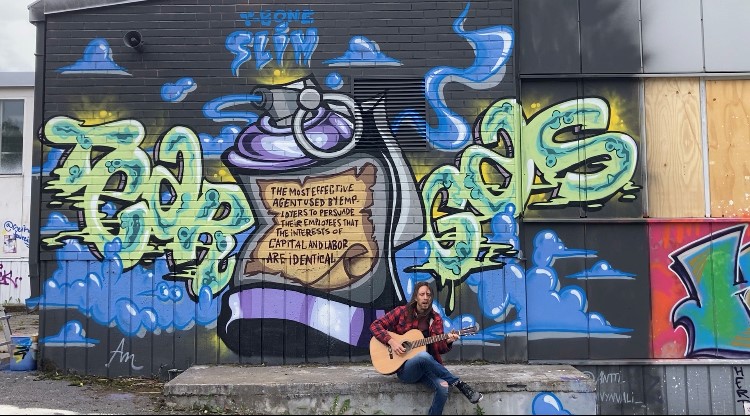 Graffiti mural on the end of a house. Text “T-Bone Slim Tear Gas” and image of a tear gas bottle with a text: “The most effective agent used by employers to persuade their employees that the interests of capital and labor are identical.” Man playing guitar in front of the graffiti mural.