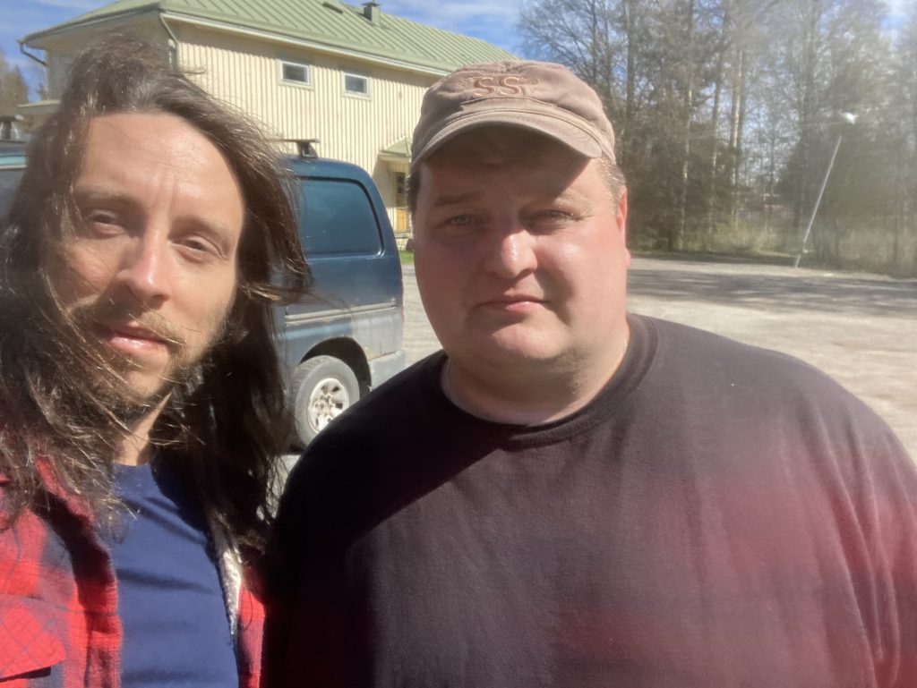 Selfie of two men standing outside. The other has beard and long hair and other a cap hat.