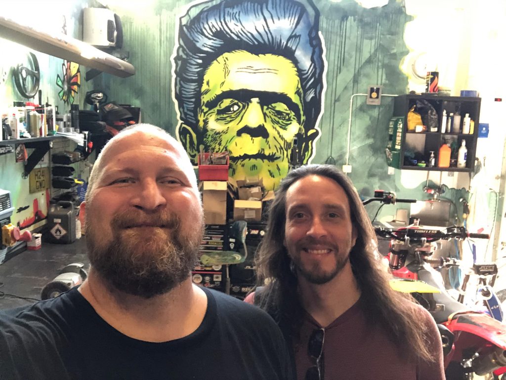 Selfie of two men in a garage. Both men have beard, the other is bald, other has long hair. Graffiti of old Frankenstein looking man in the background