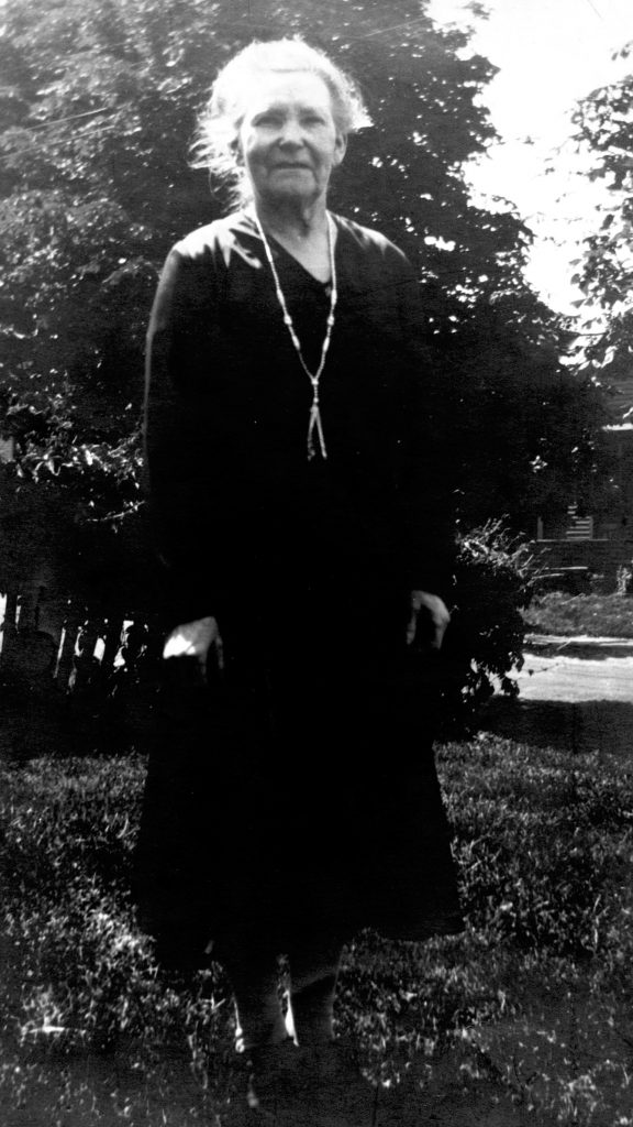 Black and white photo of an elderly lady in dark dress standing on grass by trees