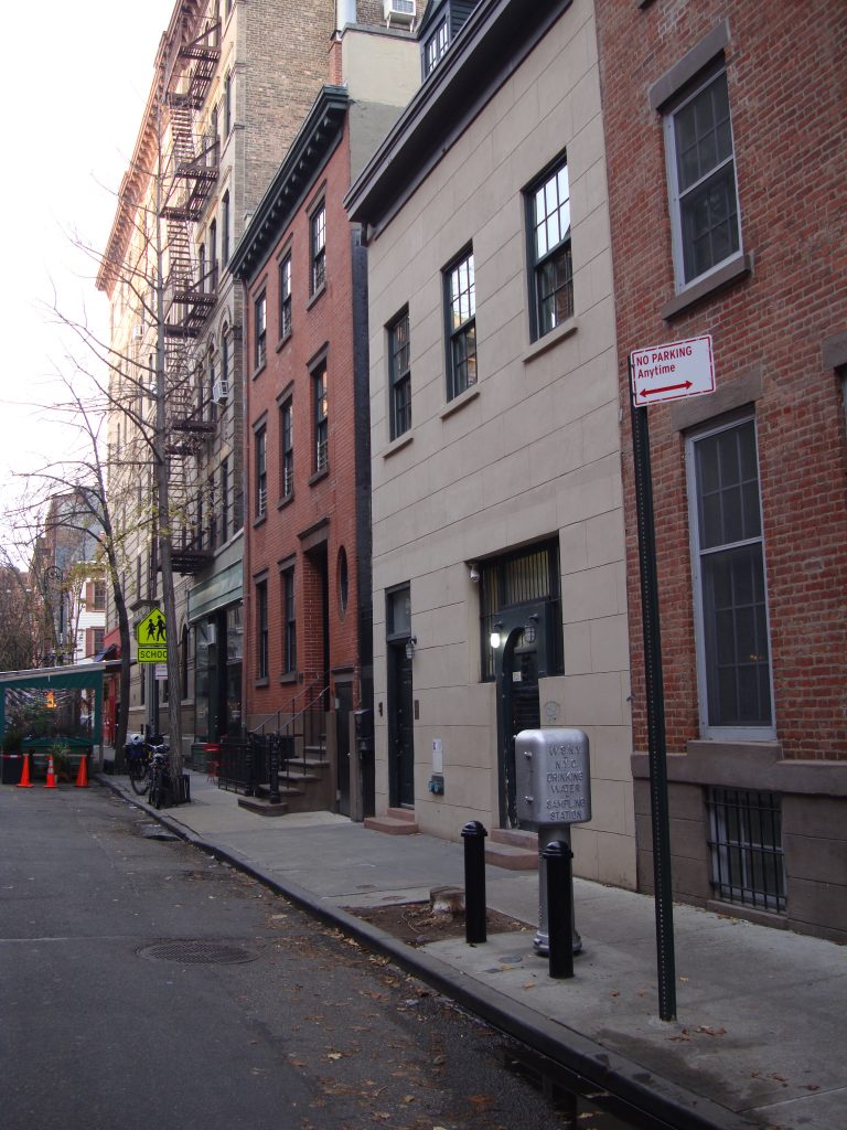 A color photograph of a typical New York street with buildings built right next to each other. In the background is a terrace extending out onto the street. Traffic signs and small trees with no leaves alongside the sidewalk.