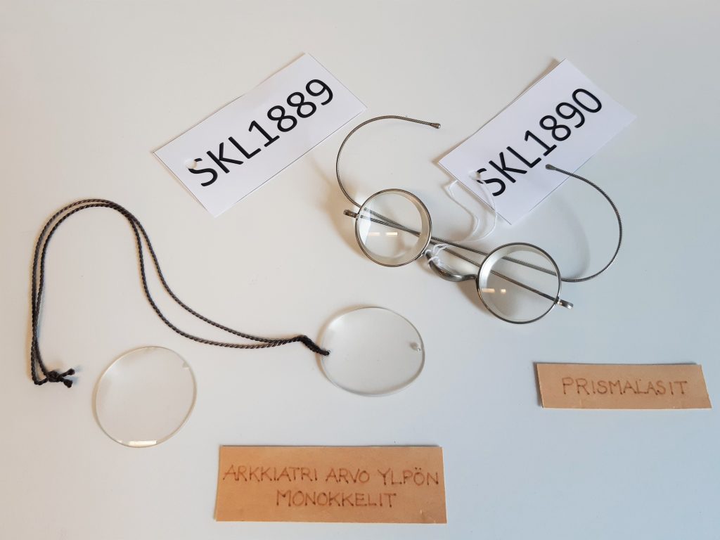 Kaksi monokkelilasia ja yhdet metallisankaiset silmälasit. Esineissä kiinni numerolaput ja niiden vieressä tekstilaput, joissa tekstit: "Arkkiatri Arvo Ylpön monokkelit" ja "Prismalasit".